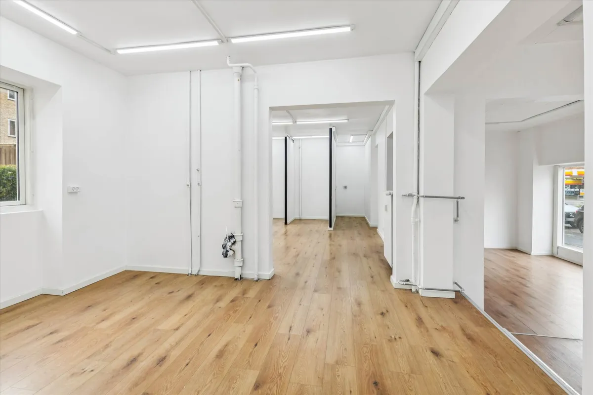 En stor, lys og tom butik med hvide vægge og gulv samt en åben dør til et andet rum med spejl.