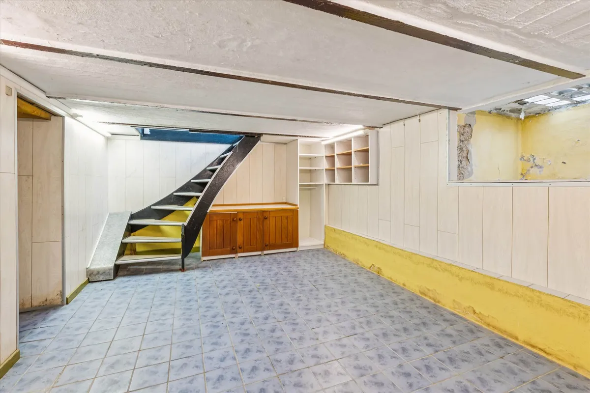 Kælder med trapper og skabe samt et hul i væggen og et vindue i loftet med lyse gulvfliser og møbler