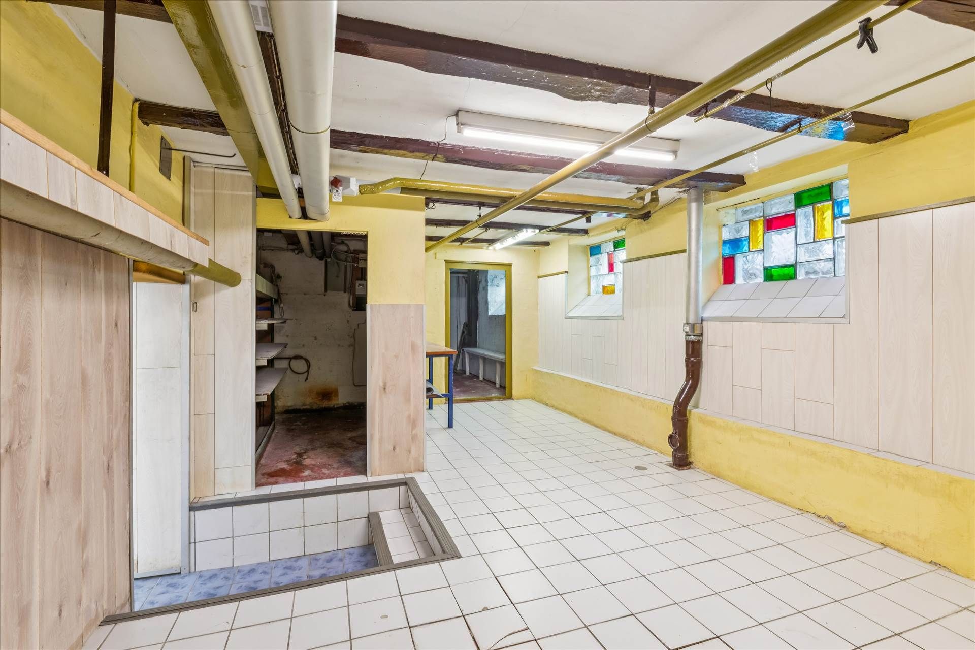 Kælder med gulvkasse og åbne døre til lagerrum og toilet i en gammel bygning med lysegul væg og gulvtæppe