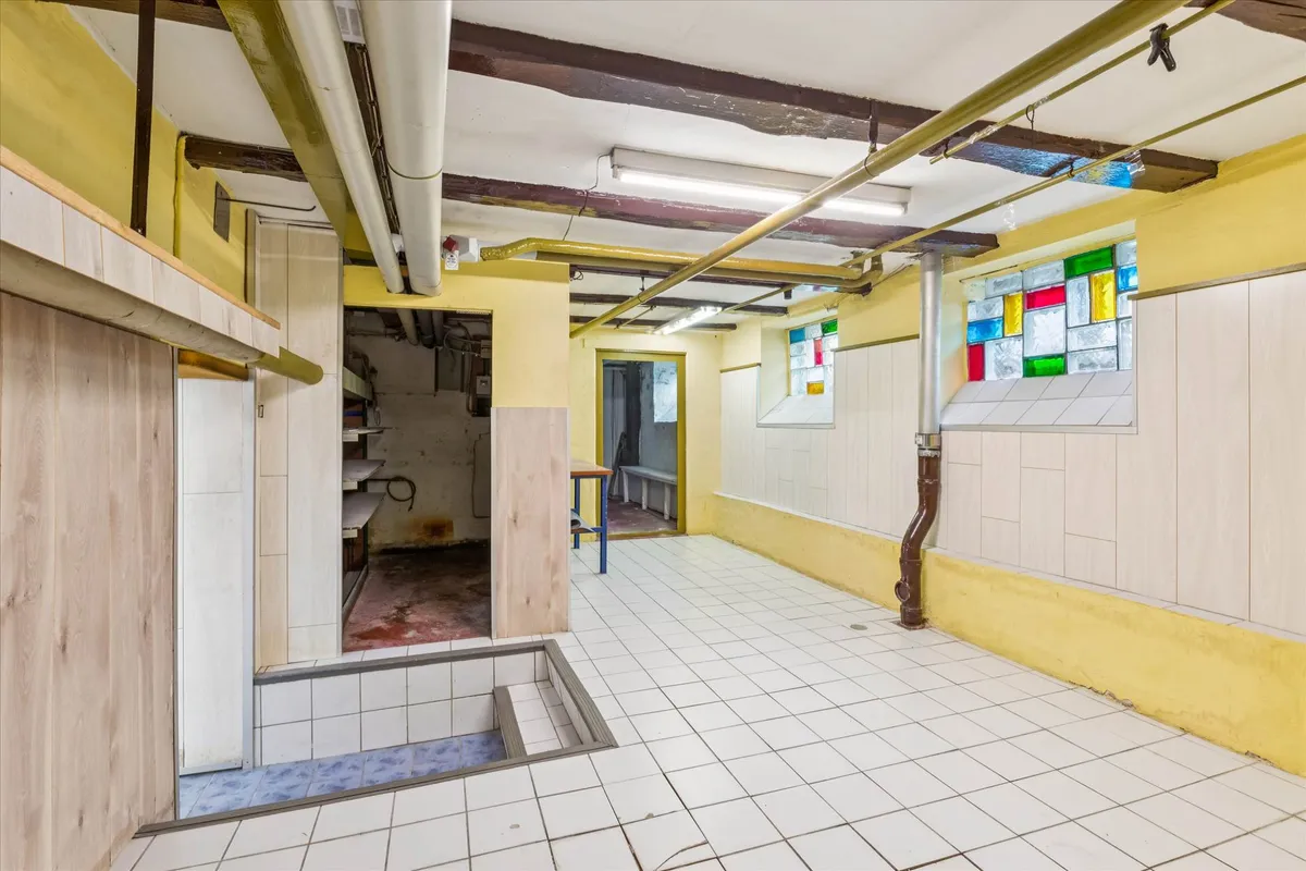 Kælder med gulvkasse og åbne døre til lagerrum og toilet i en gammel bygning med lysegul væg og gulvtæppe