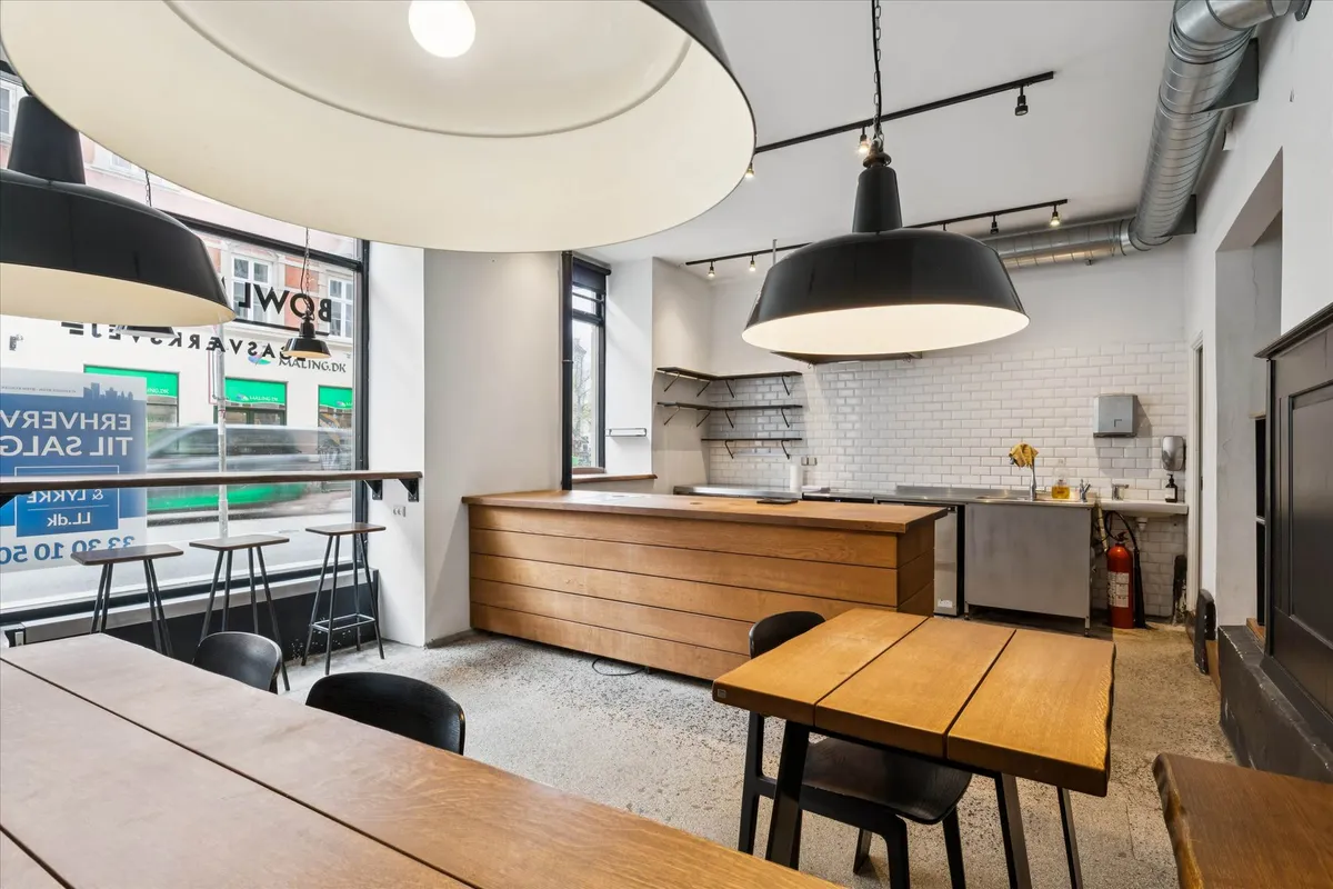 Erhvervsejendom med glasvinduer og lyse loftslamper, træborde og stole samt en åben køkkendel med højloftet