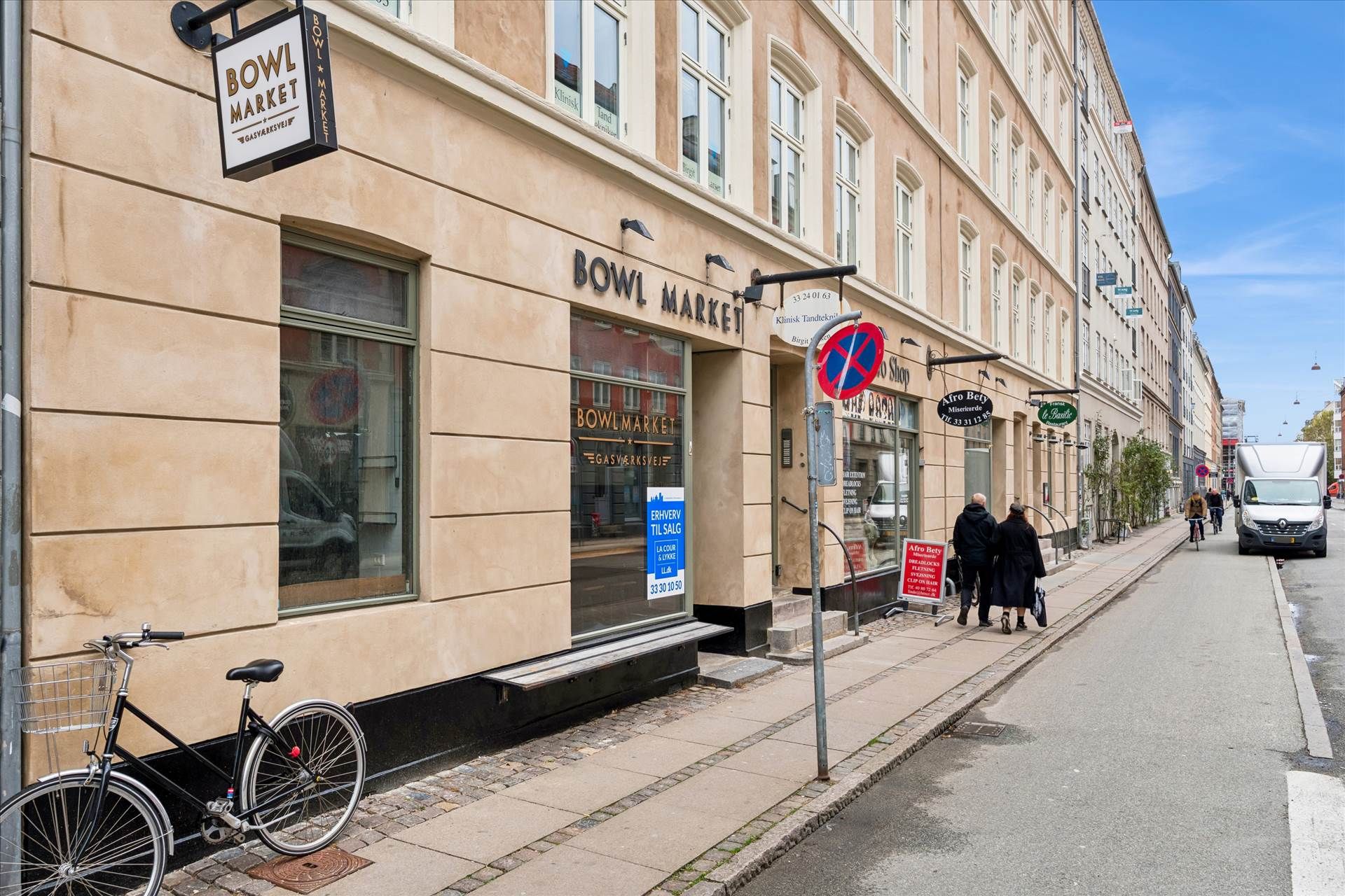 To personer går på fortovet, og der er en cykel med kurv og en bil parkeret på gaden.