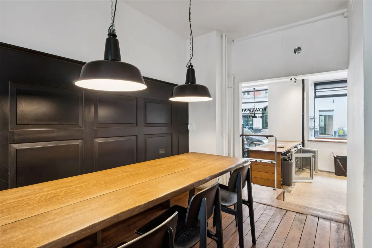 Lang bord med 4 stole og 2 lysekroner i et lokale med sort panel og en åben dør til et køkken