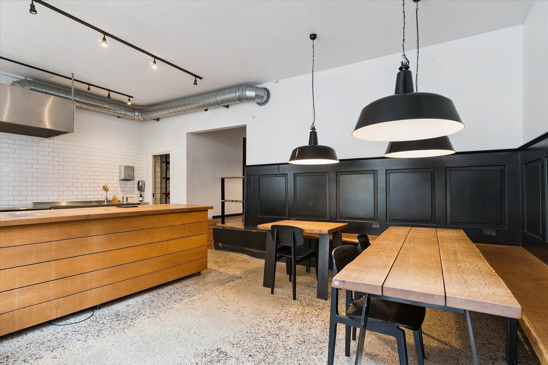 Lokale med rustfri udsugningsventilator, træborde, metalstole og hængelampe samt træskabe og hvide fliser på væggen