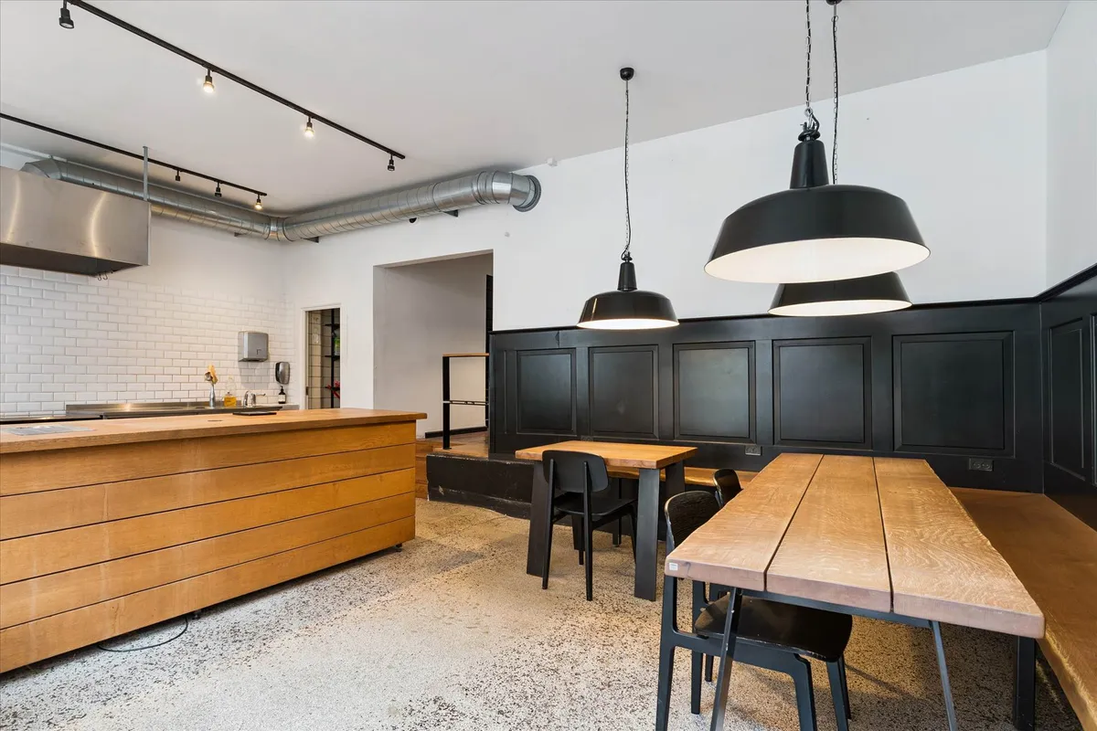 Lokale med rustfri udsugningsventilator, træborde, metalstole og hængelampe samt træskabe og hvide fliser på væggen