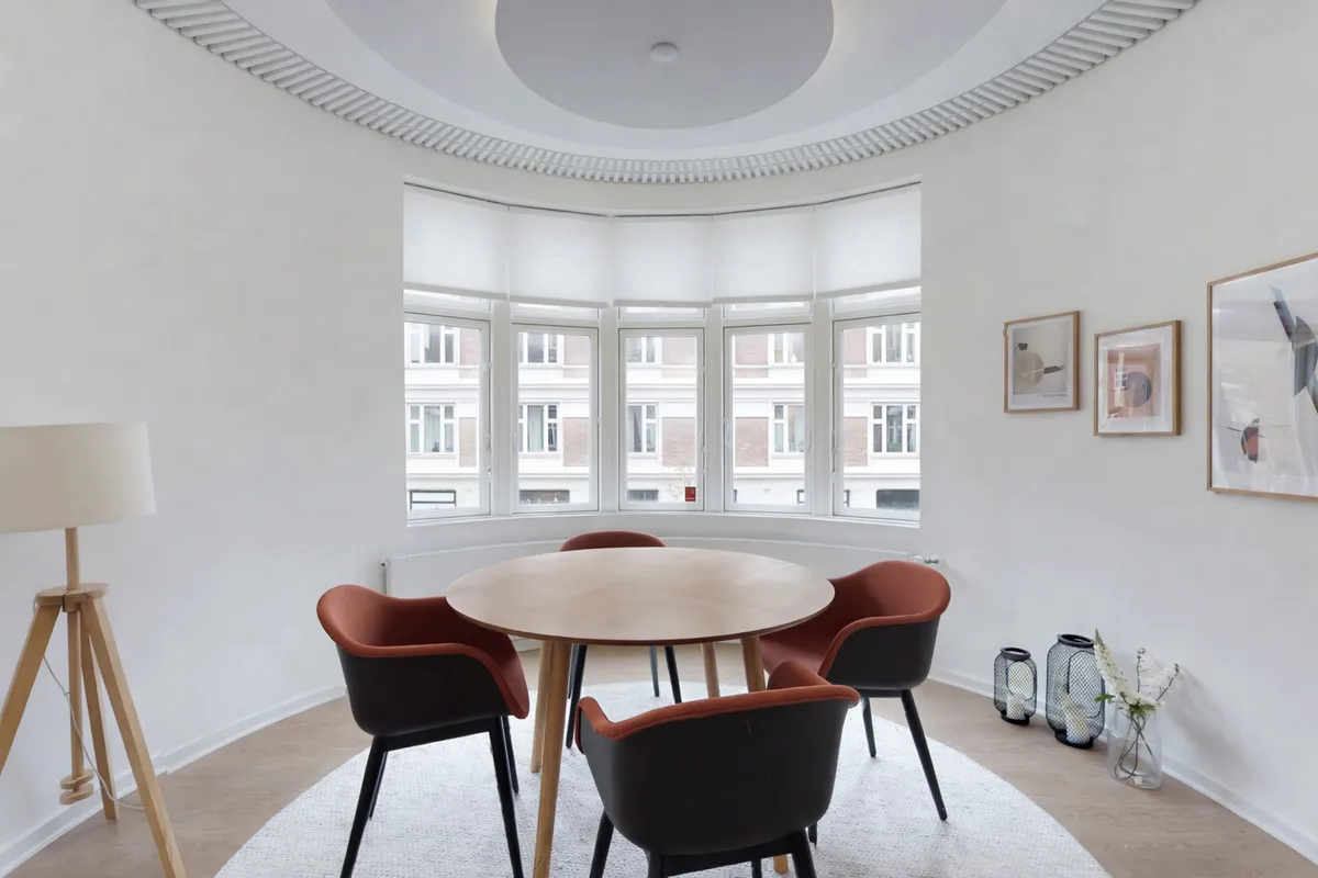 Velkommen til dette lyse erhvervslejemål med rundt bord og moderne møbler samt en hvid væg med malerier og vinduerne med udsigt til byen