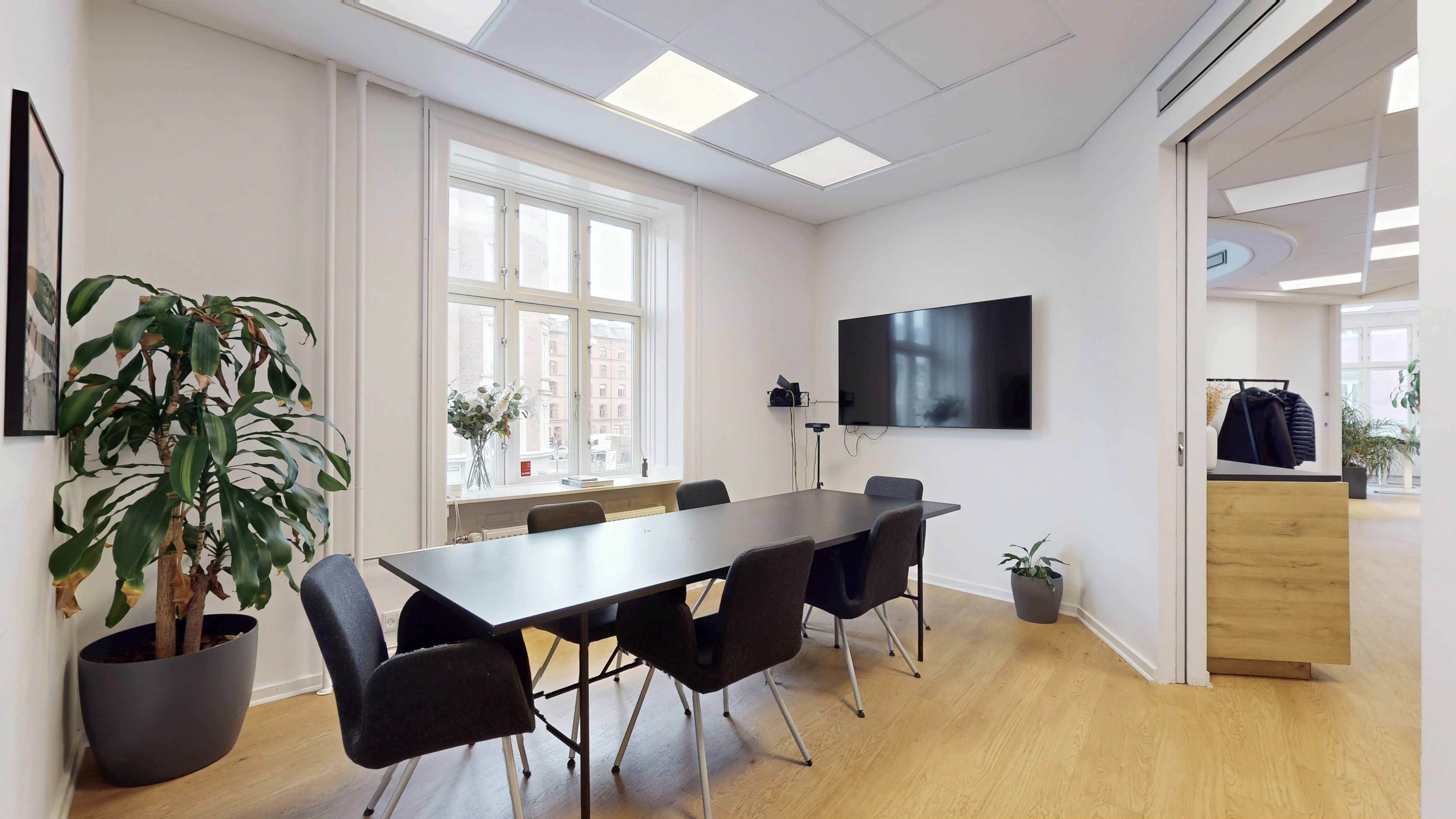 Moderne mødelokale med højt loft, lyse lamper, en stor tv-skærm, lyse møbler og store vinduer