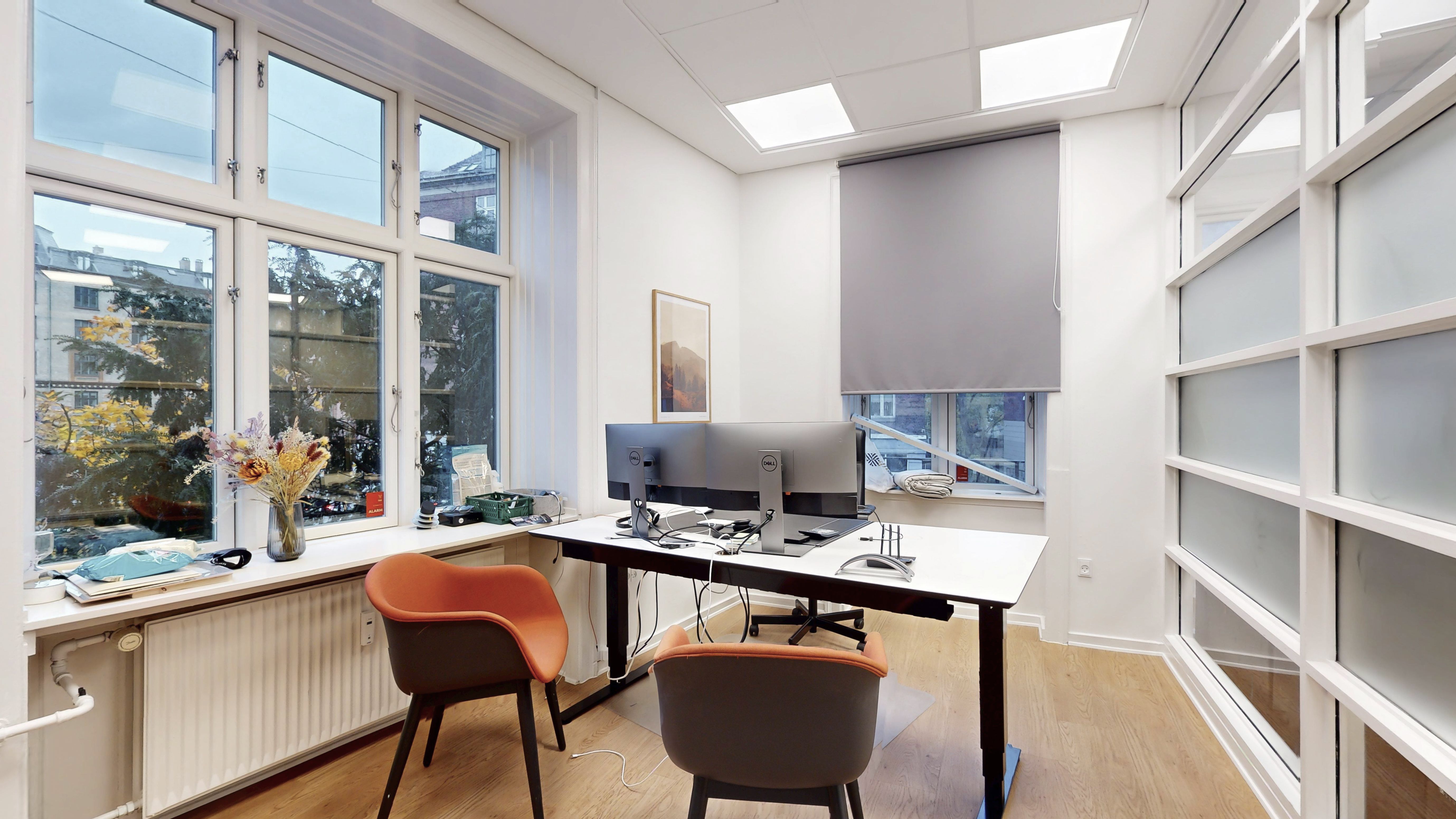 Kontor med to skriveborde og to stole samt to Dell-skærme og vinduer med gardiner og et maleri på væggen