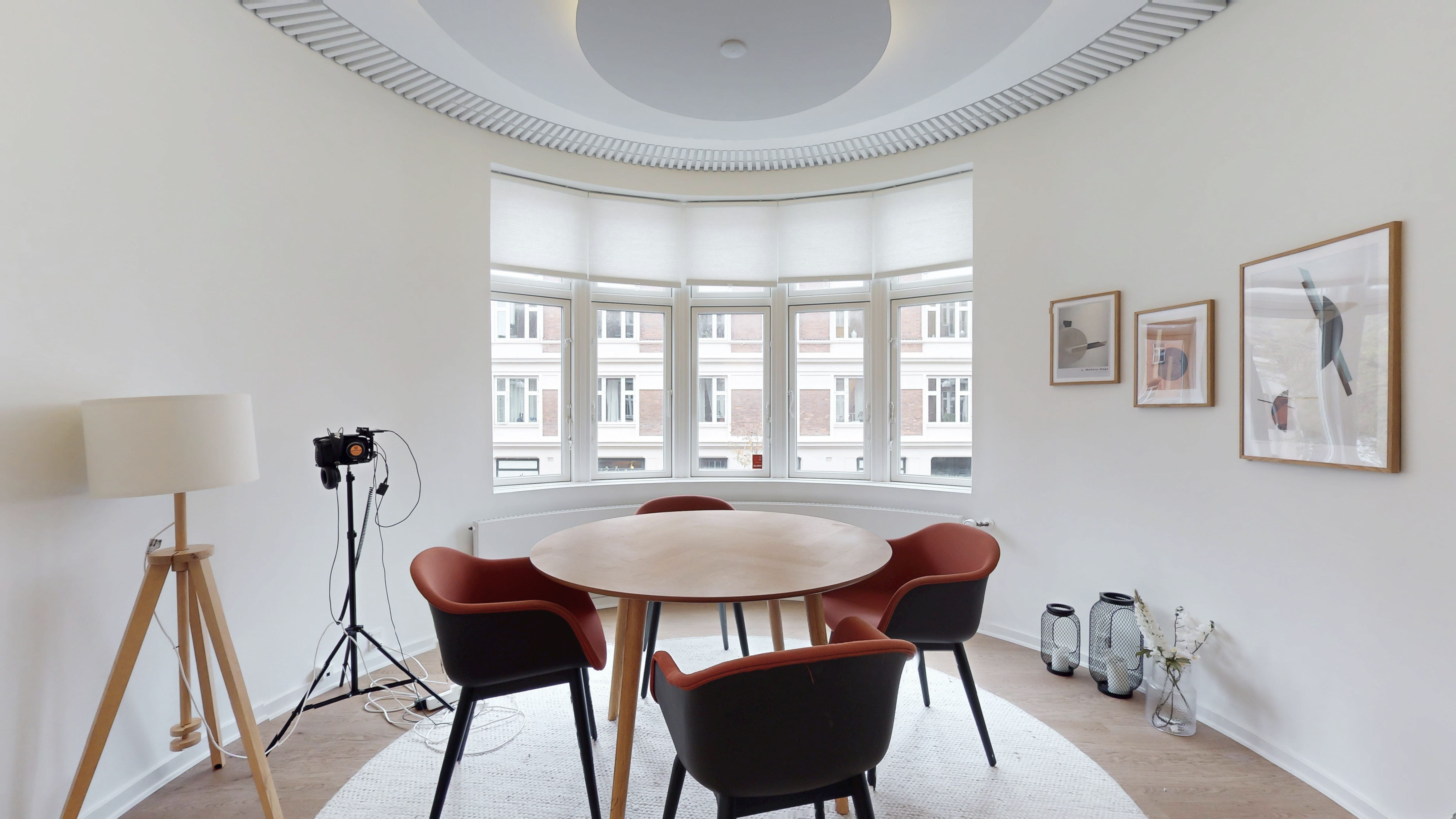 Et elegant mødelokale med rundt bord og fire stole samt et lysekron og tre vægbilleder.