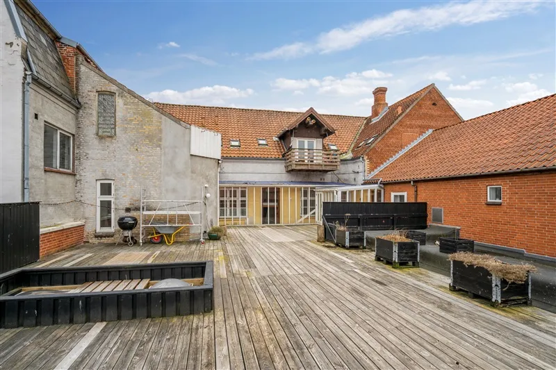 En rummelig gårdhave med trægulve, træplantekasser og et skur i baggrunden med røde tagdækninger