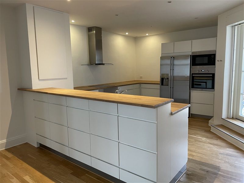 En stor hvid køkken med trægulv og moderne møbler samt hvide kabinetter og en sølvfarvet køleskab