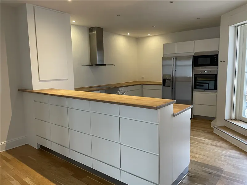 En stor hvid køkken med trægulv og moderne møbler samt hvide kabinetter og en sølvfarvet køleskab