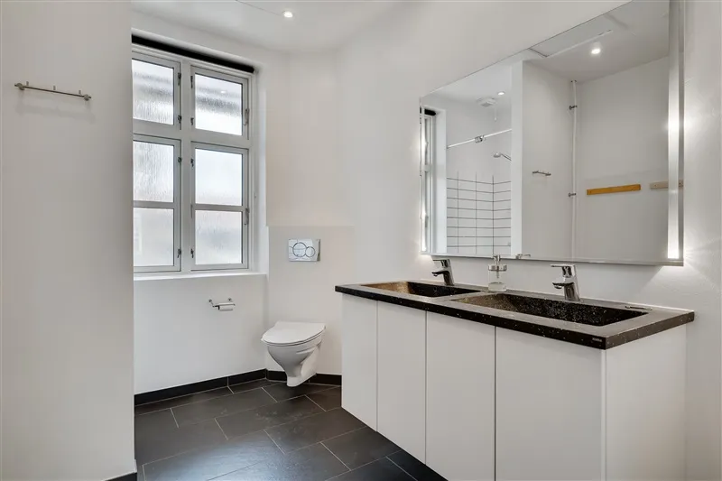 En stort badeværelse med to dobbeltvaske og et hvidt toilet med lysegrå vægge og sortgulv