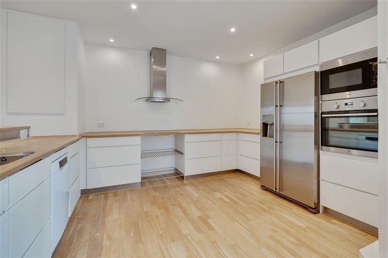 Et moderne køkken med hvide vægge, højloft, lyse trægulve og lysegrå hætte over komfuret. Hvidt køkken med lysegrå apparater