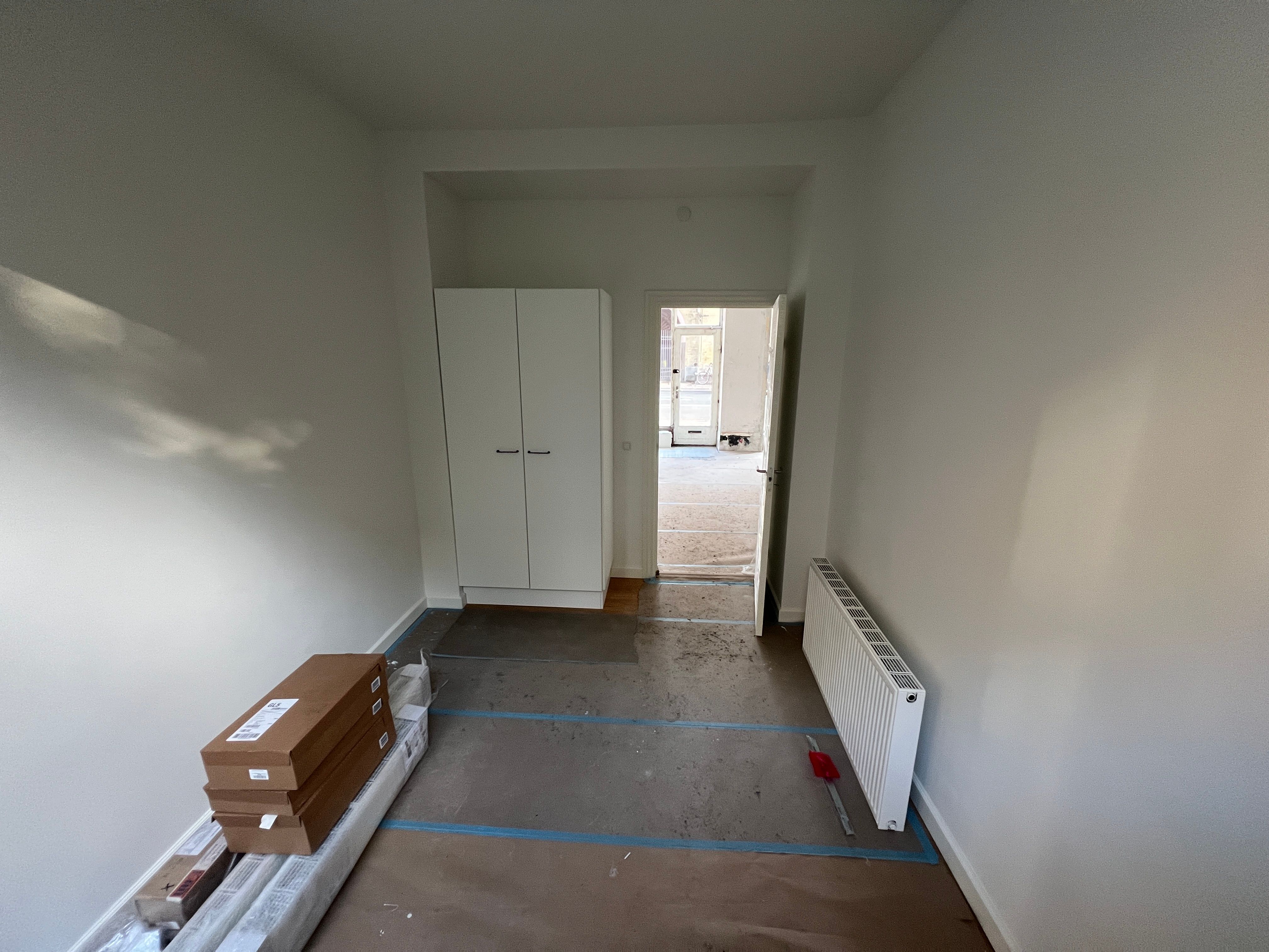 Nyrenoveret tomt rum med hvide vægge og gulvtæppe, og en hvid garderobe mod væggen, tomme bokse og en gulvmåler