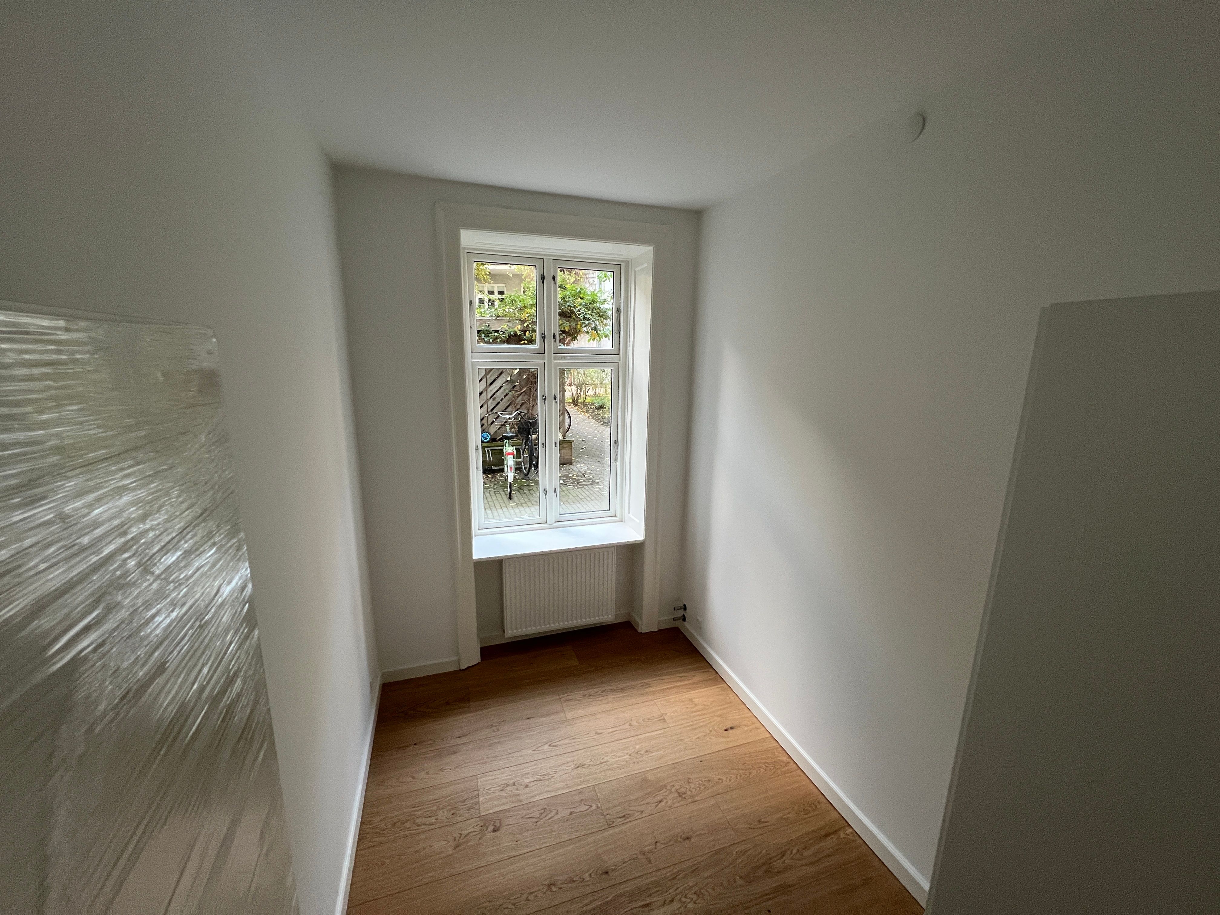 Kort og smal korridor med hvide vægge, hårdt trægulv og enkeltvindue med sølvfarvet ramme