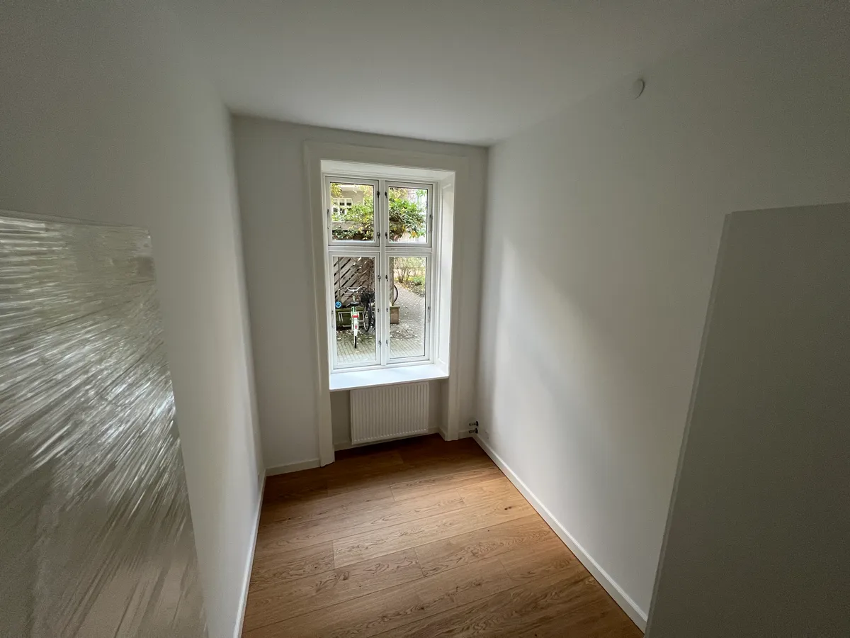 Kort og smal korridor med hvide vægge, hårdt trægulv og enkeltvindue med sølvfarvet ramme