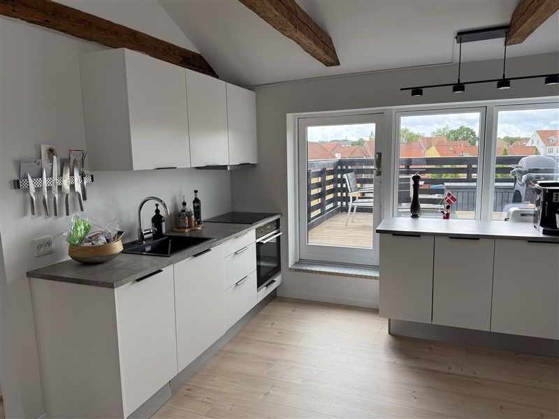 Køkken med hvide loftsbjælker og skur, moderne udformning med hvide skabe og en stor balkon med udsigt til bygninger og