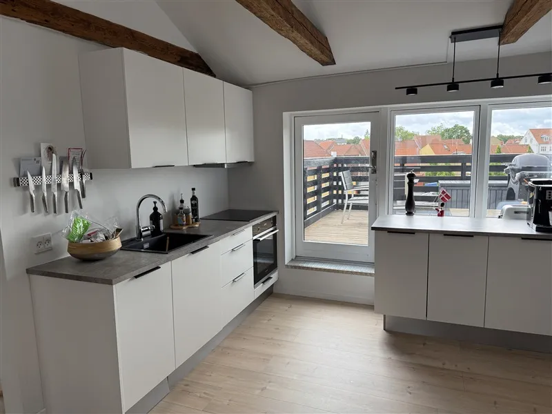 Køkken med hvide loftsbjælker og skur, moderne udformning med hvide skabe og en stor balkon med udsigt til bygninger og