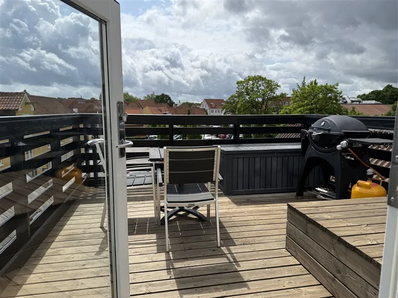 Balkon med moderne møbler og grill samt udsigt over byen med grønne træer og regnfulde skyer