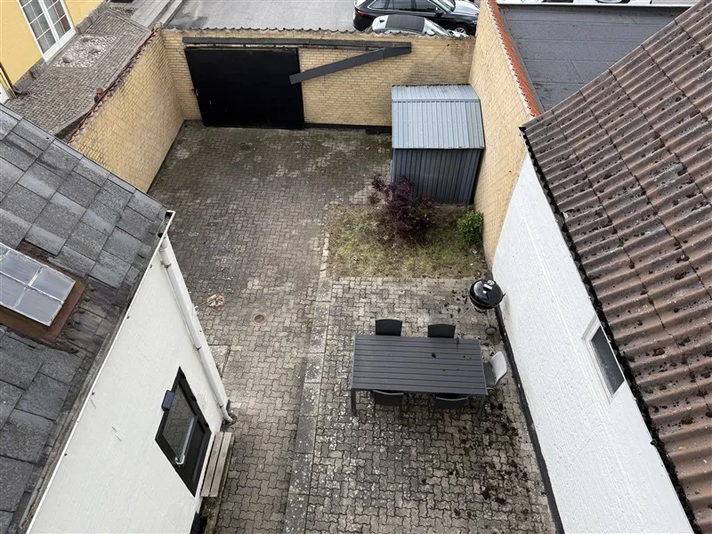 Et hus med en stenlægget have og en udendørs spisebord med fire stole omkring. Have er omgivet af huse og er placeret nær en