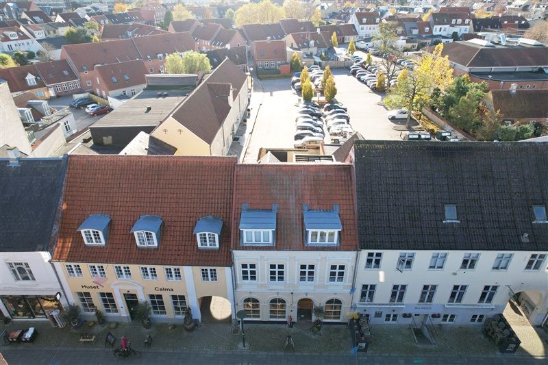 Middelfart by med rækkehus og butikker. Biler parkeret og gågade. Solskin og træer. Forretningsvinduer med skilte.