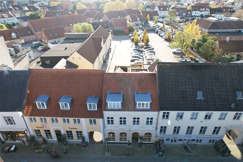 Middelfart by med rækkehus og butikker. Biler parkeret og gågade. Solskin og træer. Forretningsvinduer med skilte.