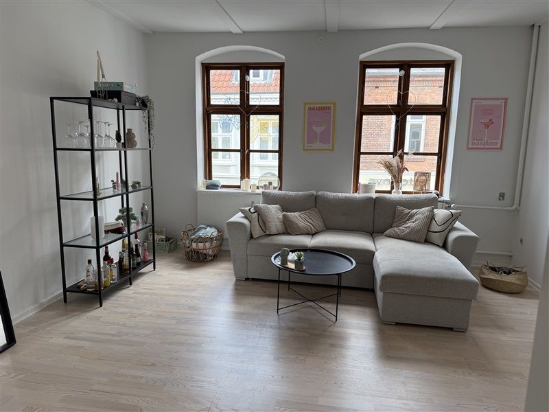 Et moderne stue med trægulv og en stor L-formet sektionssofa med puder, to vindu med buede ruder, en ståbord med en plante