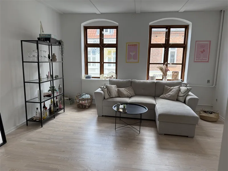 Et moderne stue med trægulv og en stor L-formet sektionssofa med puder, to vindu med buede ruder, en ståbord med en plante