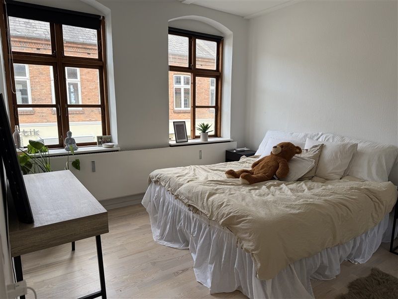 Et soveværelse med hvidt loft og vægge, trægulv, og to store vinduer med udsigt til en murstensbygning, og et brun