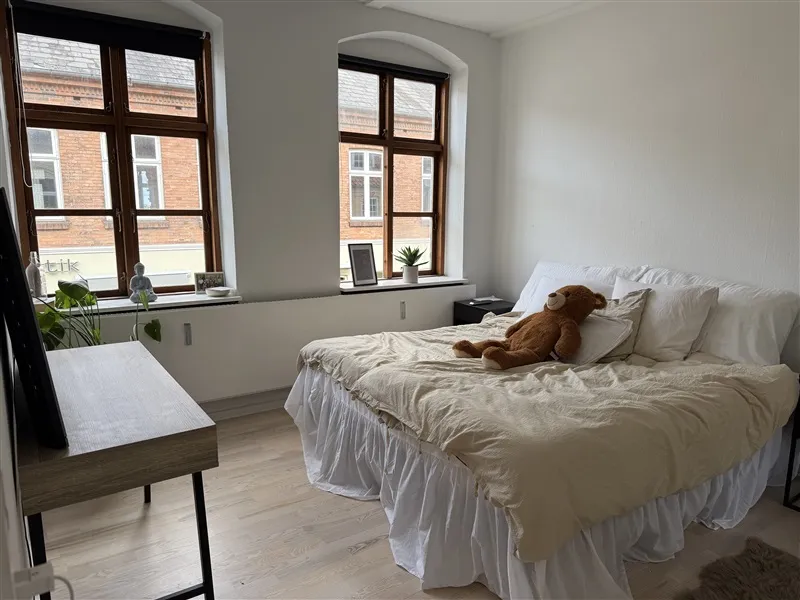 Et soveværelse med hvidt loft og vægge, trægulv, og to store vinduer med udsigt til en murstensbygning, og et brun