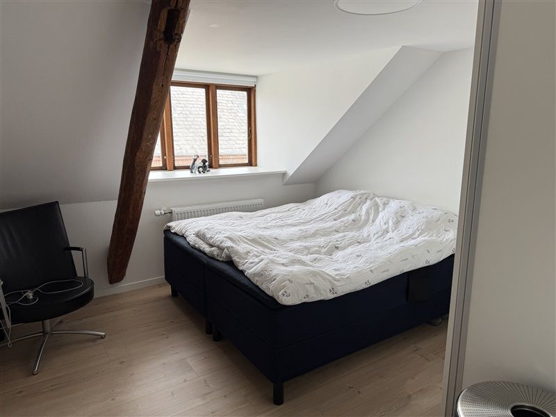 Et soveværelse med en dobbeltseng, hvidt sengelinned og en træbjælke ved siden af vinduet