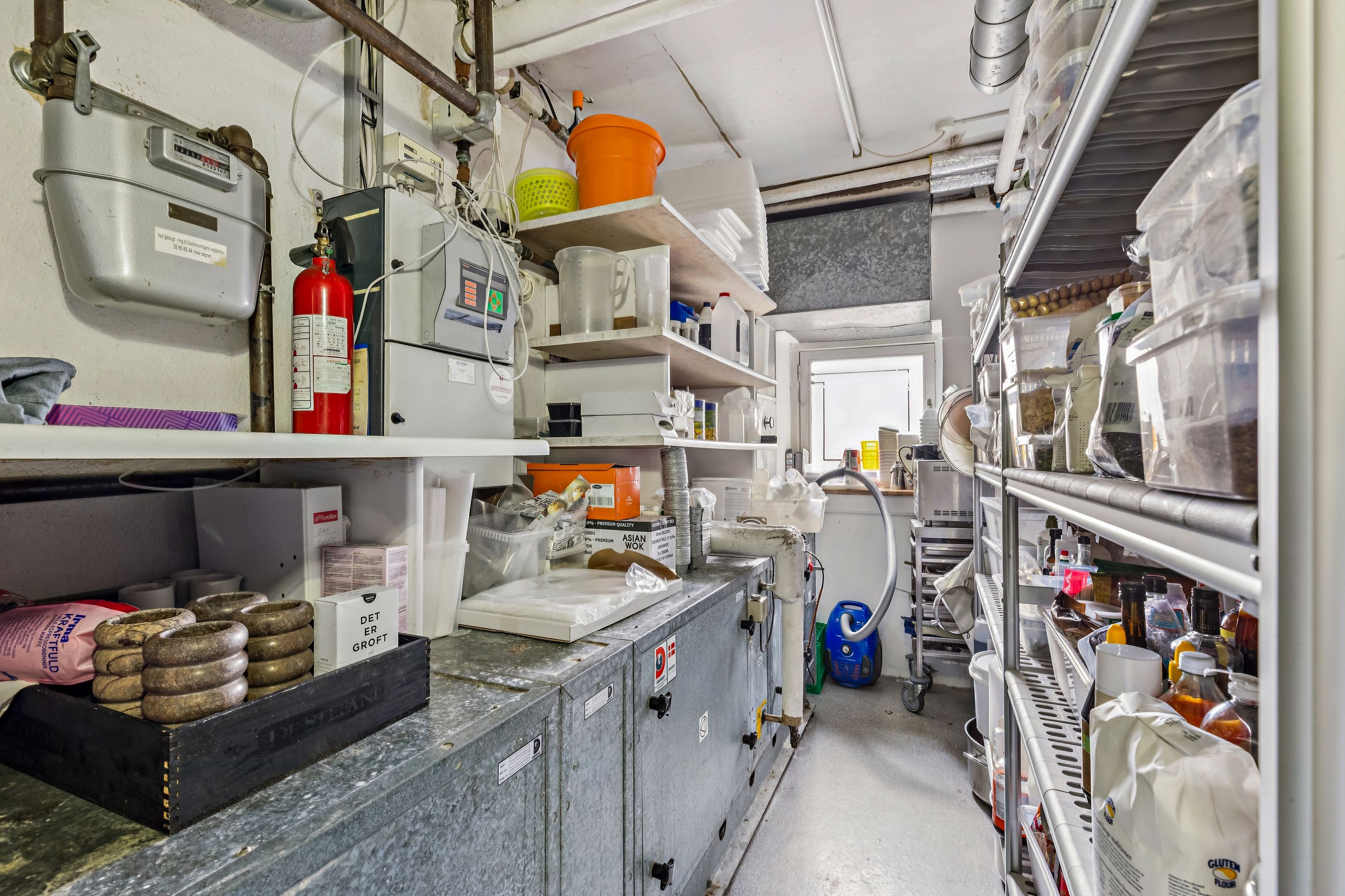 En lejlighed med lagerrum med høje hylder, kasseapparater, flasker og andet lagermateriale samt en rødt brandvæsen