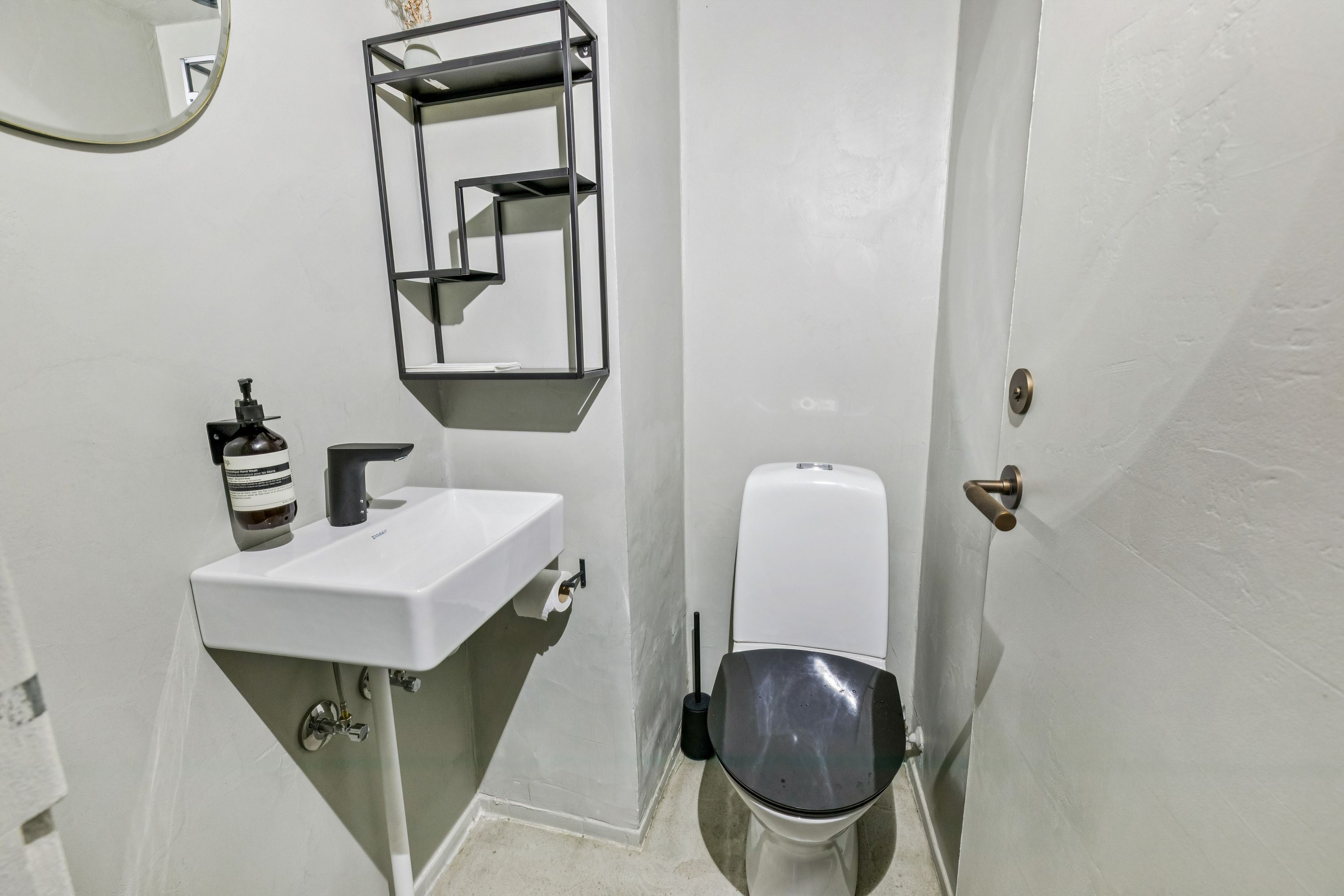 Et lille badeværelse med hvidt toilet, hvidt vasketøjsskab og sort sædeskærm og sæde. Der er en hvid sæbe dispenser og et