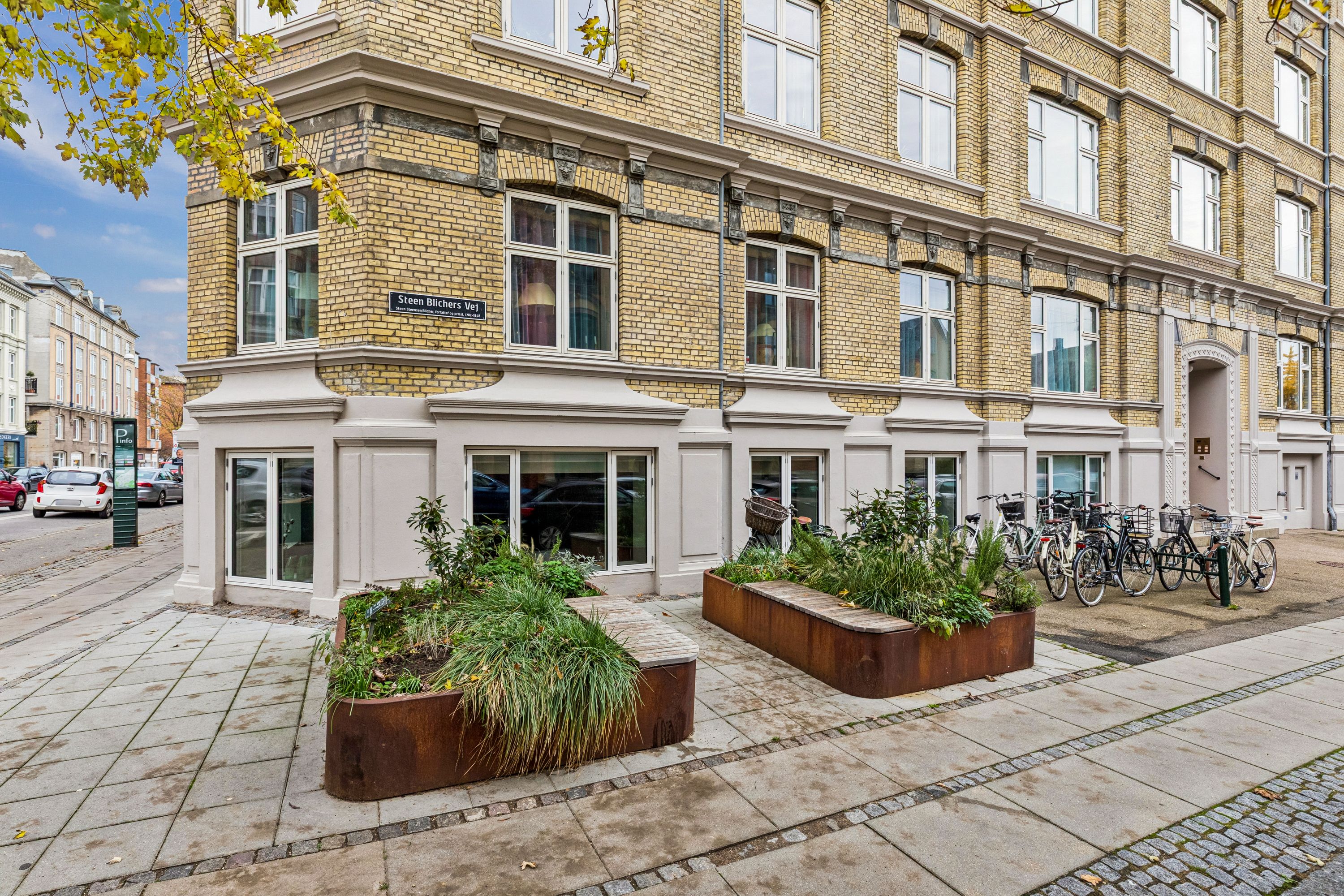 En bygning med flere lejligheder, store ruder og gårdhave med planter og cykler i nærheden
