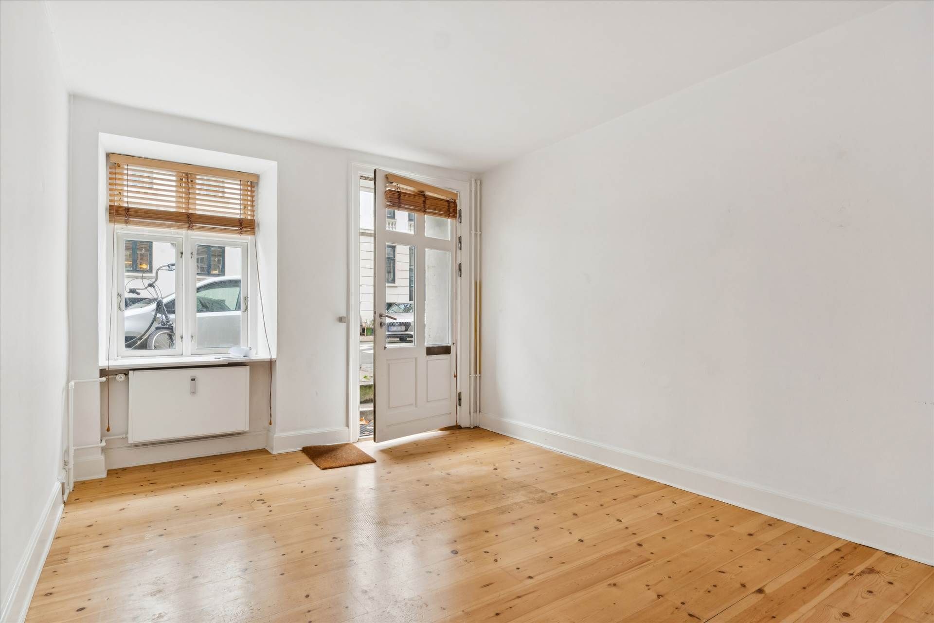 Lys, tom stue med hvide vægge og trægulv, åbent hvidt dørkarm og vindue med lysegardiner og udsigt til parkeringsplads og