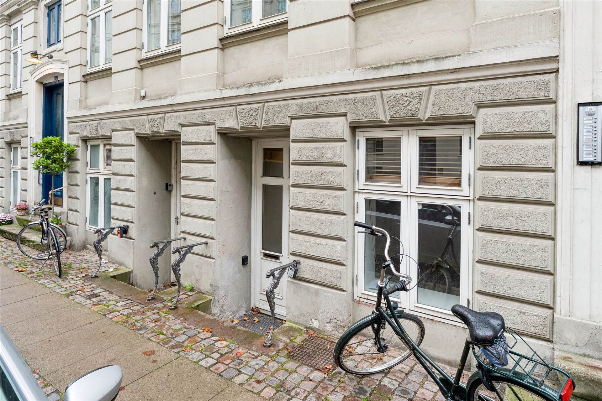 To cykler er parkeret udenfor en bygning med vinduer og døre, en af dørene er åben og en cykel har en sæk i bagenden