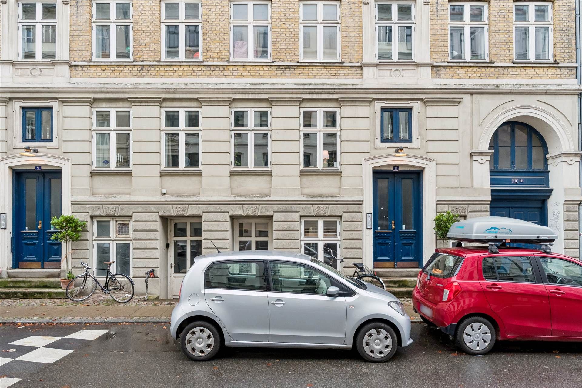 To små biler parkeret i København med lyserøde og lyseblå vinduer og højt gulv. En cykel parkeret foran døren og et hus med
