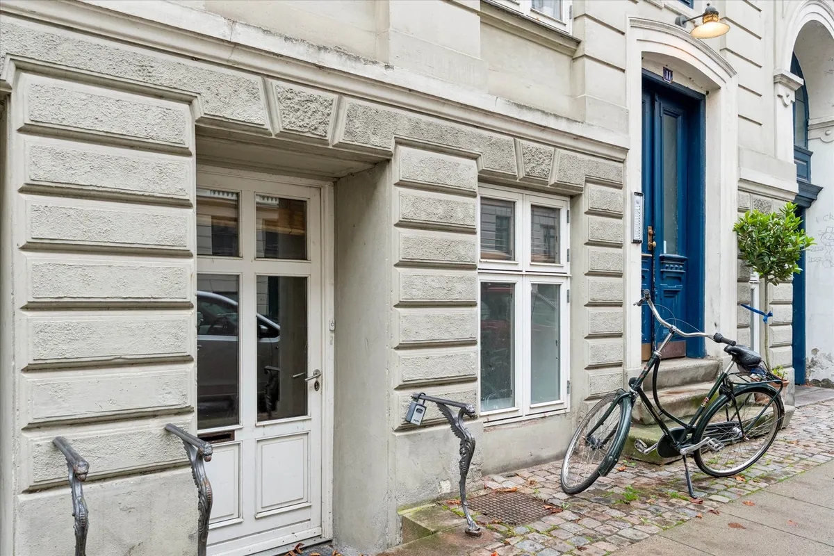 En grøn cykel parkeret på brolægget udenfor en hvid bygning med glasruder og en slank søjle med en lampeskærm.