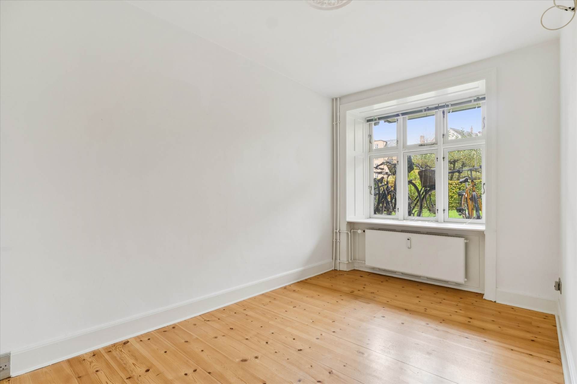 Et tomt værelse med hvide vægge og trægulv, med et vindue med trækarmer og hvide ruder og en radiator under vindueskarm