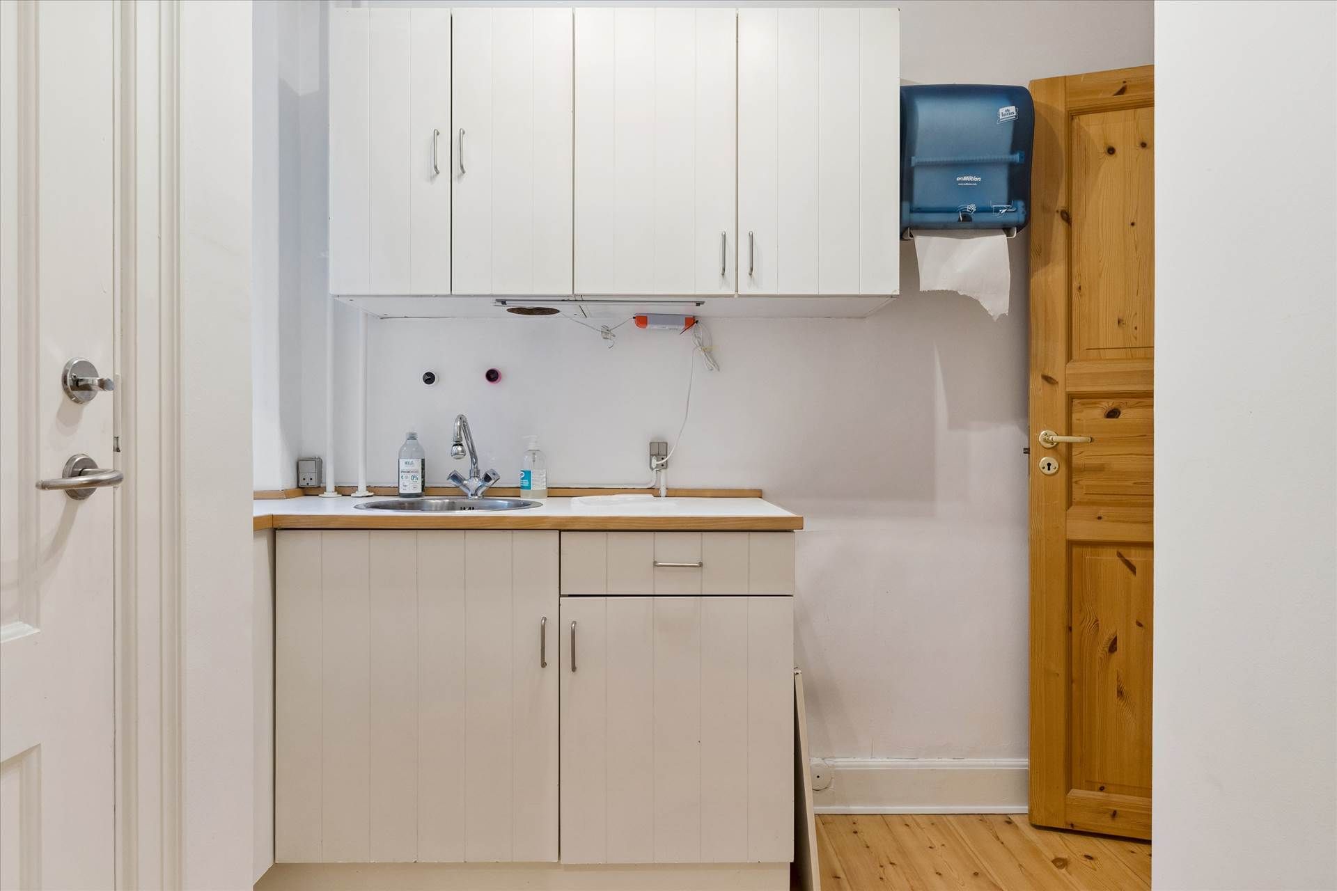 En lille køkken med vask, håndklædeholder og hånddesinfektionsmiddelflaske samt et toiletpapirautomat og åbent træskab