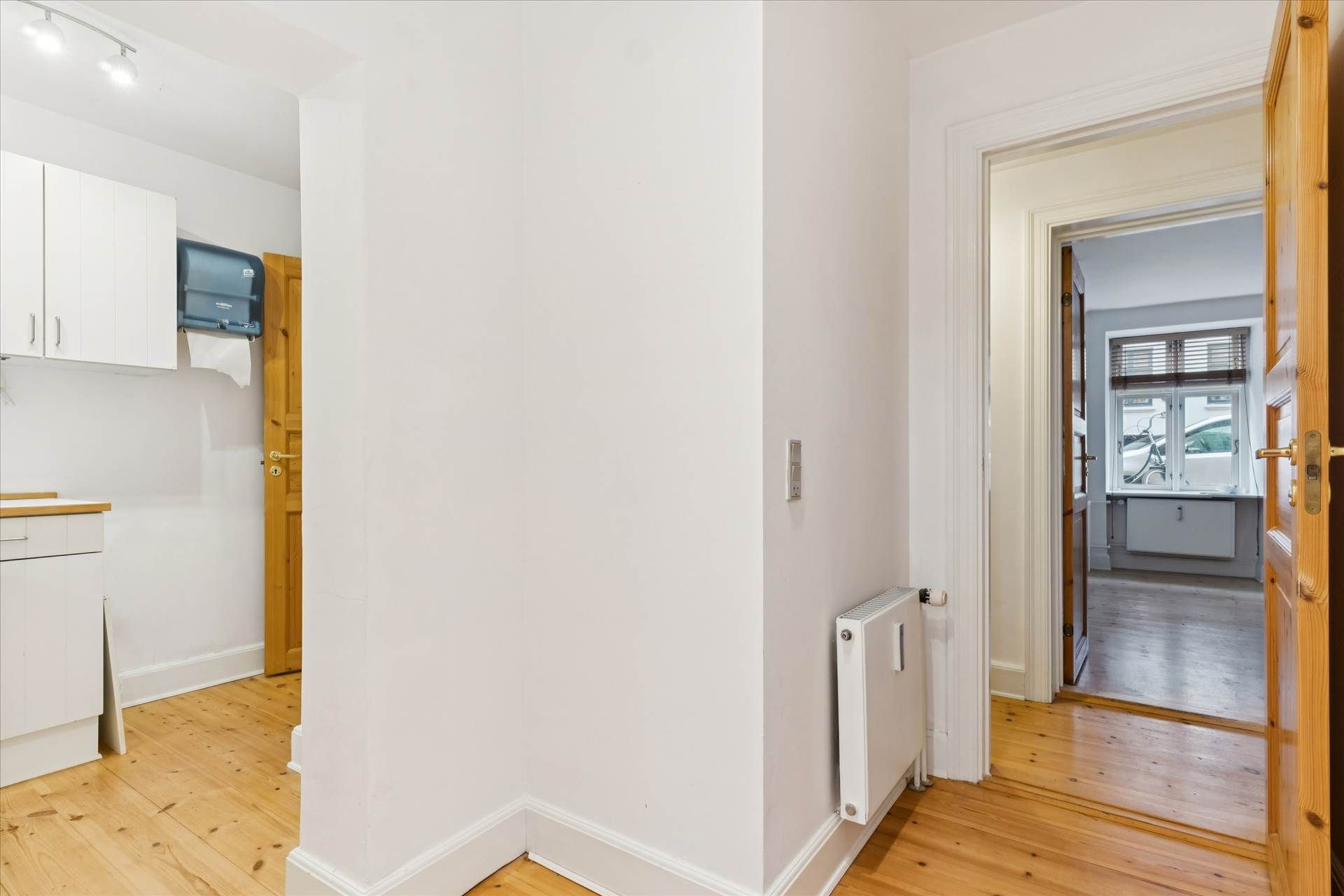En indre gang med lyse trægulve og hvide vægge, åbne døre, en radiator og en toiletpapirholder på en højre hængeskab