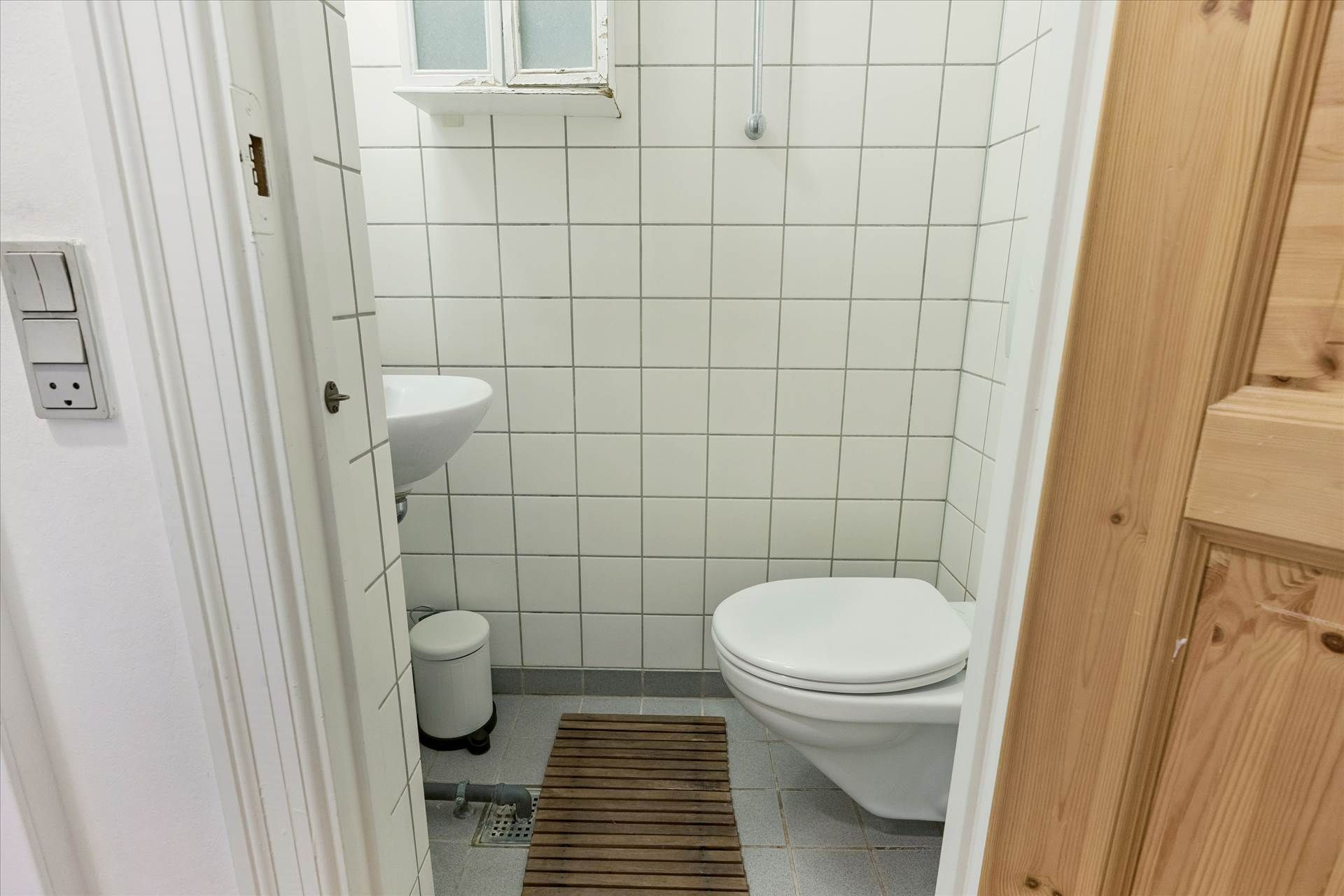 Et lille toilet med toilet, håndvask og tøjskab samt en udtræksbar tørretumbler. Der er et trægulv og hvide kakeloverflader