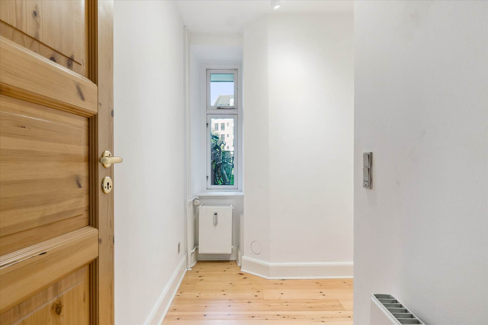 En trædør og en radiator i en hall med hvidt panel og gulvtæppe og et vindue med udsigt til en bygning