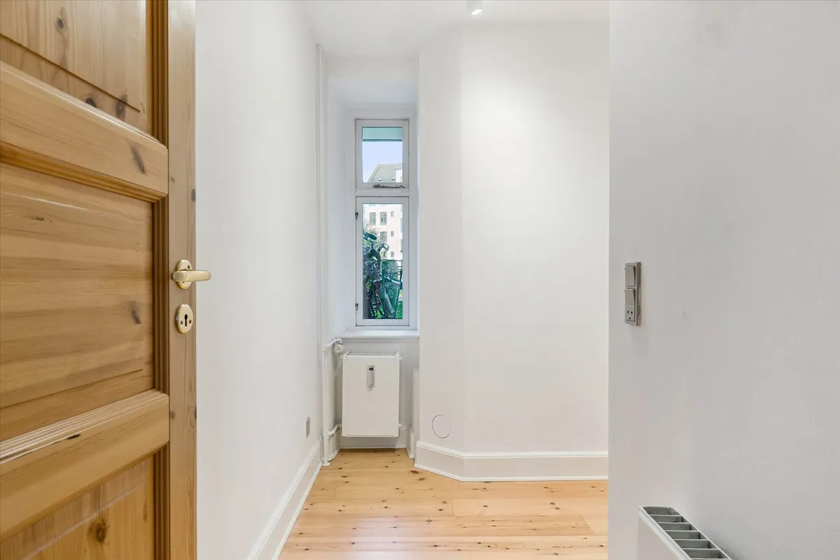 En trædør og en radiator i en hall med hvidt panel og gulvtæppe og et vindue med udsigt til en bygning