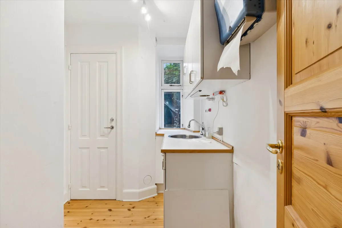 Minimalistisk indrettet køkken med hvide vægge, lysekroner, lille vask, køkkenhylder og toiletpapirhylde samt vinduesparti