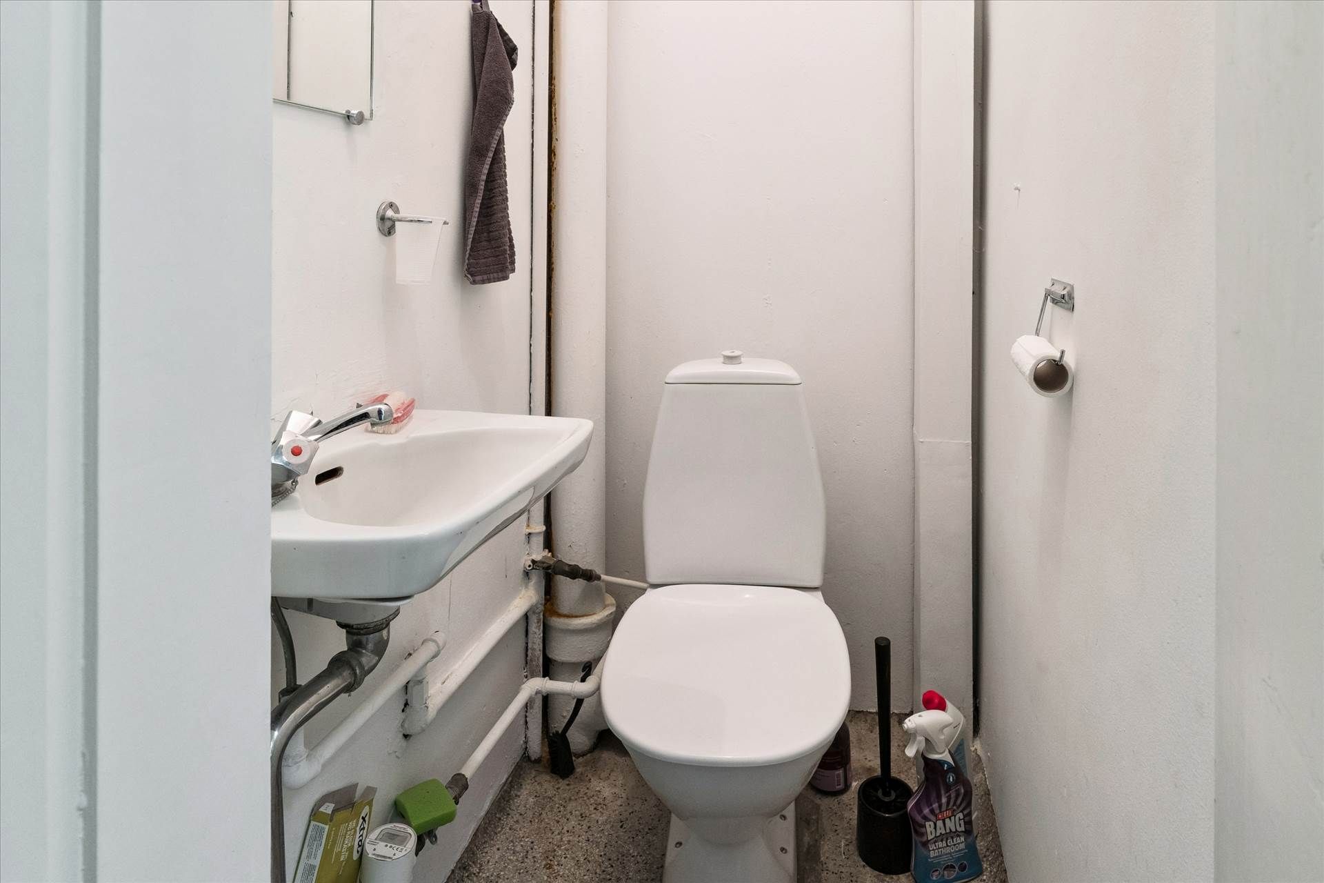 En lille toiletrum med hvidt toilet og hvidt vask. Der er tørklæde og toiletpapir. Der er rengøringsmiddel i nærheden af