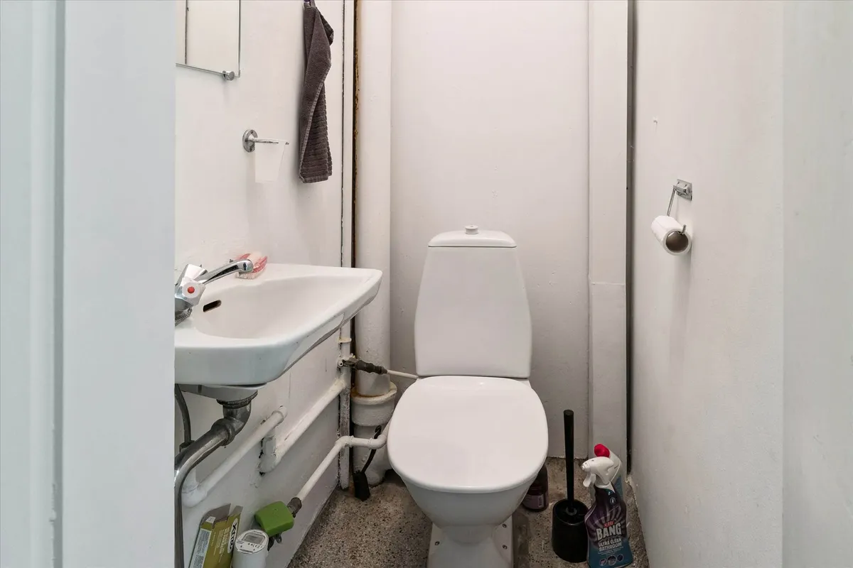 En lille toiletrum med hvidt toilet og hvidt vask. Der er tørklæde og toiletpapir. Der er rengøringsmiddel i nærheden af