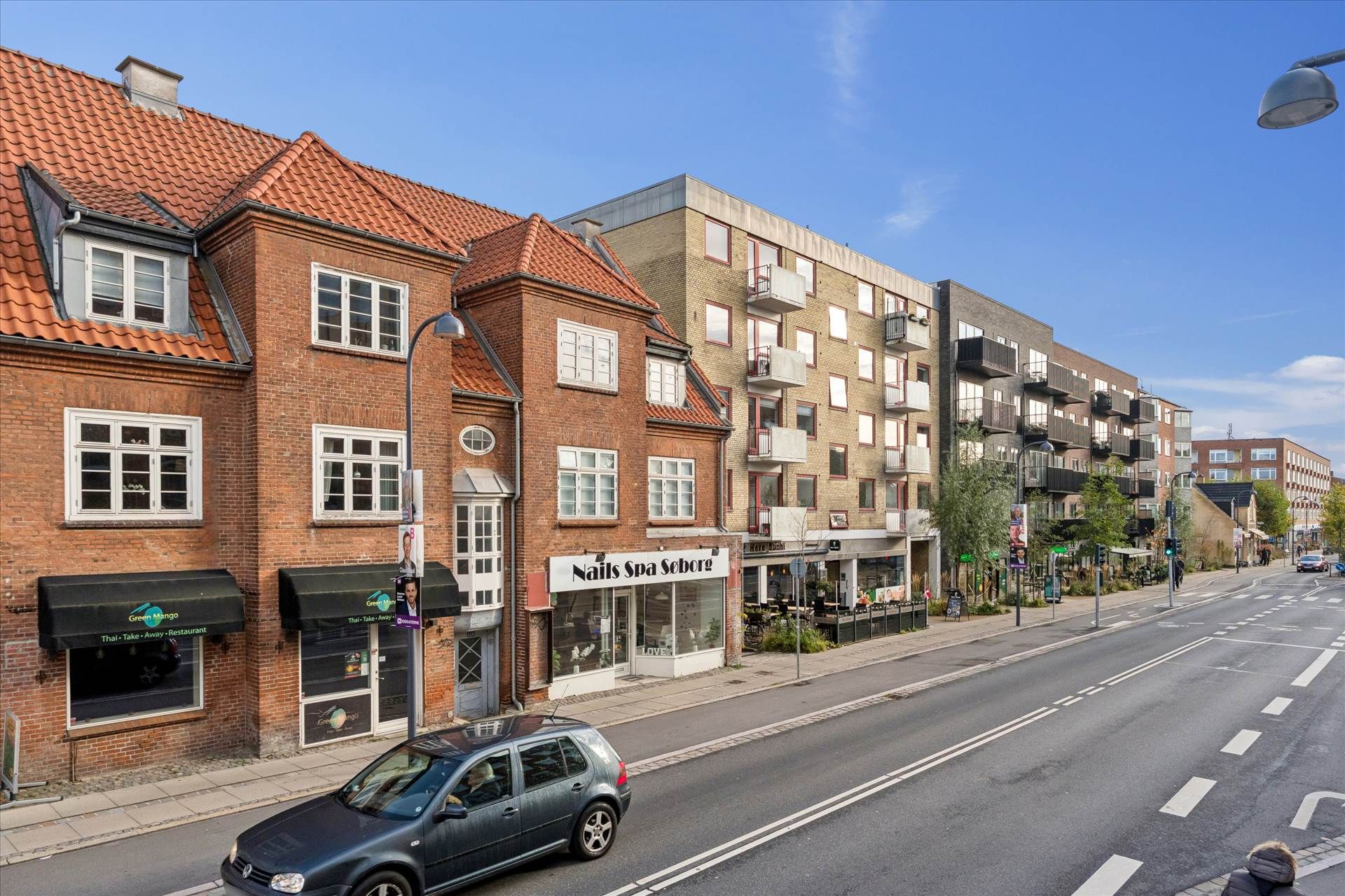En bil kører på Søborg Hovedgade, forbi butikker og huse med rød tegltag og balkoner