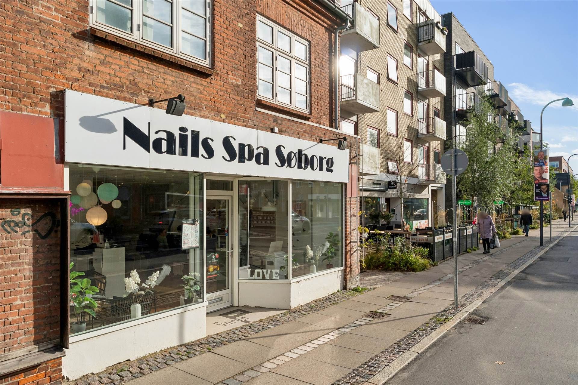 Butik med navnet Nails Spa Søborg ligger på en travl gade med fortov og grønne planter